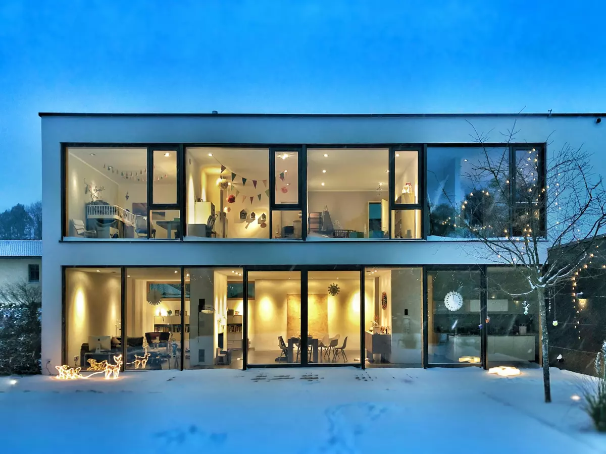 A modern house with large windows showcasing the interior, taken at dusk with the exterior lights on, showing a cozy interior and a snowy garden. 